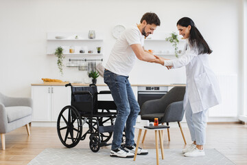 Full length view of attractive Caucasian woman helping young patient with disability to stand from wheelchair in modern light kitchen. Medical specialist supporting patient in living room of home.