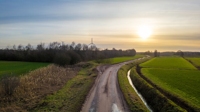 This landscape image offers a view of a winding country road at sunset. The road curves around a lush green field and a row of bushes, guiding the eye towards a solitary wind turbine in the distance
