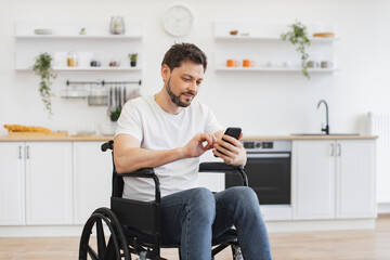 Smiling Caucasian adult in wheelchair looking at cell phone screen while having rest in comfortable...