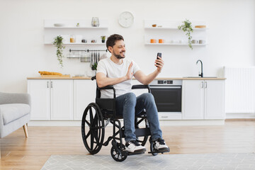 Cheerful wheelchair user in casual outfit utilizing phone webcam while enjoying free time. Bearded...