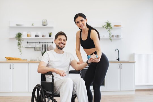 Portrait of handicapped mature man training with his wife or female trainer using dumbbells. Young physiotherapist or wife in sportswear helping Caucasian male patient in wheelchair exercise at home.
