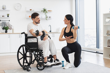 Obraz premium Rehab fitness for young male patient. Millennial physiotherapist in sportswear helping adult man with a disability in wheelchair to workout with dumbbells, to recover from injury at home.