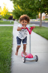 Cute, smiling, young diverse boy riding his scooter on the sidewalk on a fun summer day outdoors in his neighborhood. Lifestyle photo for you diverse child. Safe Neighborhood concept photo