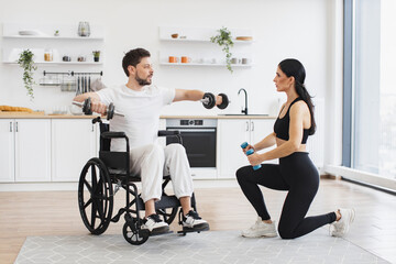 Female physiotherapist or wife showing mature male patient in wheelchair how to exercise with...
