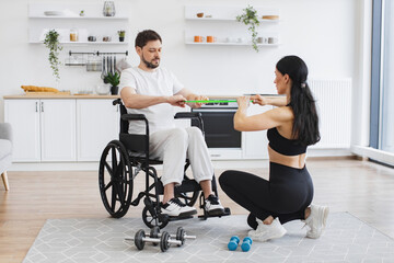 Young physiotherapist or wife in sportswear helping male patient in wheelchair exercise at home. Concept of rehabilitation of disabled people. Handicapped mature man training with stretching bands .