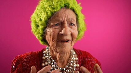 Closeup of funny confused, elderly woman, 80s, do not understand, wearing green wig or hat isolated on pink background. Concept of youthful old female.