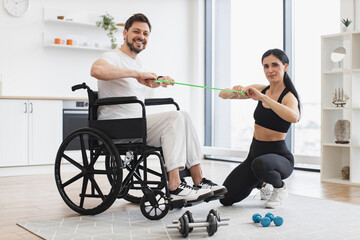 Female physiotherapist showing adult man in wheelchair how do exercises with stretching bands at...