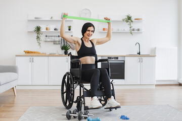 Full length view of Caucasian female with mobility impairment doing seated exercises with stretching bands in open-plan kitchen. Positive woman in sportswear gaining health benefits from training.