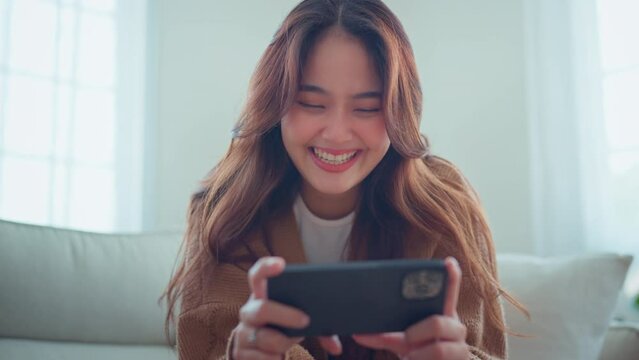 Happy young asian woman surfing social media on mobile phone, enjoying amazing news , Female having fun playing games chatting in social media sit on couch at home, watching relax movie