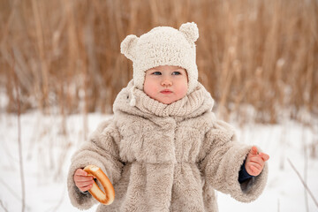 A cute little kid in a plush hat and a fur coat has a steering wheel on the background of a snowy winter landscape with reeds. The winter season, a cozy childhood. Russian Slavic style.