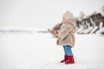 Cute little kid in a plush hat and fur coat eats a bagel on the background of a snowy winter...