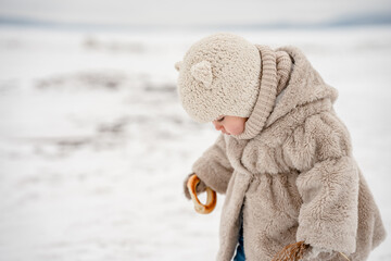 Cute little kid in a plush hat and fur coat eats a bagel on the background of a snowy winter...
