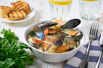 Creamy soup with mussels and shrimps served with lemon and parsley on a white table.