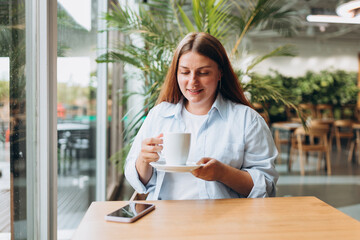Happy woman wear blue shirt hold cup drink coffee sit alone at table in coffee shop, cafe or...