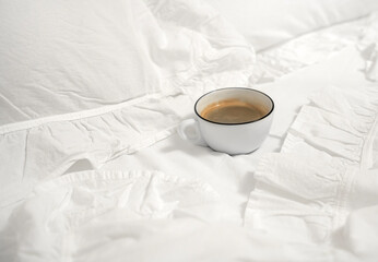 A white cup of cappuccino stands on a bed in white linen, a pillow and a blanket
