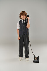 well-dressed young gentleman in elegant vest with tie and glasses holding retro telephone on grey