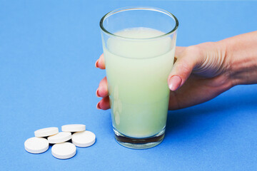 Dissolving medicine in water. Hand holding glass of fizzy water with soluble tablet inside. Pile of white pills isolated on blue screen.