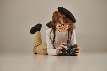 young photographer in beret and glasses taking photo on camera and lying on floor, creative boy