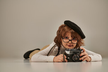 happy young photographer in beret and suspenders taking photo on retro camera and lying on floor