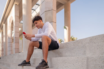 A sporty man sits typing a text message on his cell phone.