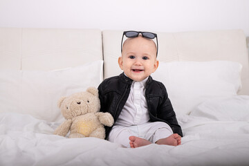 Portrait of smiling stylish child sitting on bed in white clothes wearing leather jacket with teddy bear in hands and glasses on head european child