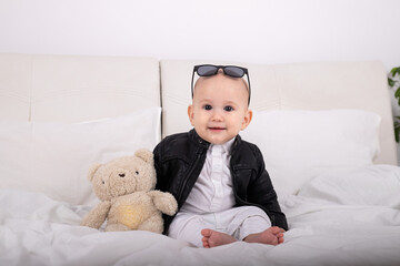 boy sitting on bed in white clothes leather jacket with teddy bear in hands with glasses on head on valentine's day newborn baby infant boy gentleman