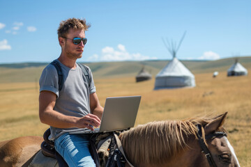 A young digital nomad wearing sunglasses sits on a horse with his laptop in a field, traditional yurts visible in the background under a clear blue sky. - obrazy, fototapety, plakaty
