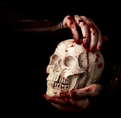 Naklejka premium Bloody hands of a witch holding human skull over black background