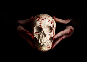 Bloody hands of a witch holding human skull over black background