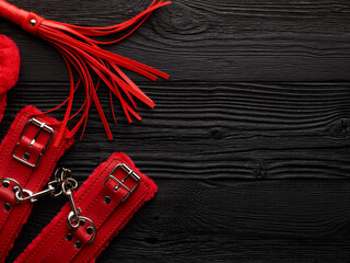 Bright red handcuffs and red whip over black wooden background