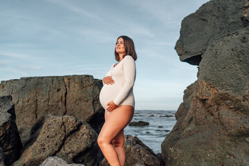 pregnant woman in bodysuit stands proudly on a rocky beach, her hand cradling her full belly,...