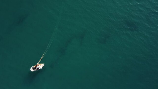 drone, vista dall'alto. Pescatore su piccola barca in mare limpido raccoglie rete da Pesca. Movimento a salire.