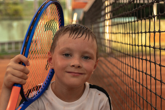 Portrait of a young tennis player sitting at the net of a tennis court with a racket in his hands. Boy tired after tennis training.Active children concept