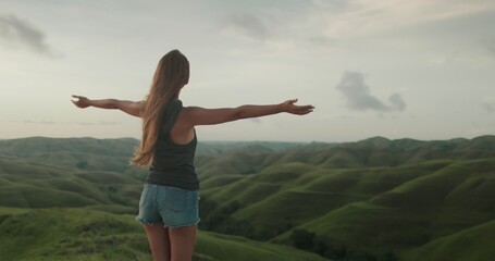 Woman enjoy sunset landscape, raises hands against green hills valley. Long hair brunette girl...