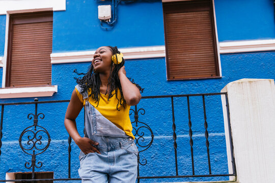 African Latina lady with braids feeling the music in yellow headphones, standing by a blue house, eyes closed with joy.