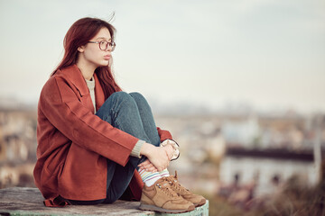 Pensive teenage girl in glasses with long red hair in red coat sits with his knees bent and looks...