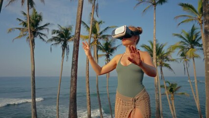 Woman in VR headset explores virtual realm among palm trees by ocean. Tech enthusiast experiences immersive reality, gestures in tropical paradise at sunset, virtual travel trend concept.