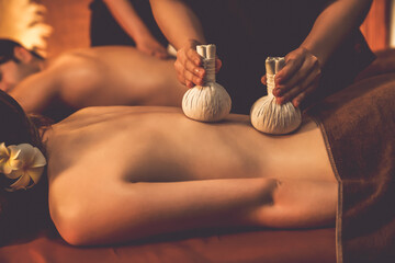 Hot herbal ball spa massage body treatment, masseur gently compresses herb bag on couple customer body. Serenity of aromatherapy recreation in warm lighting of candles at spa salon. Quiescent - obrazy, fototapety, plakaty