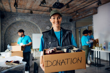 Happy female volunteer with carboard box of donated clothes at community center looking at camera.