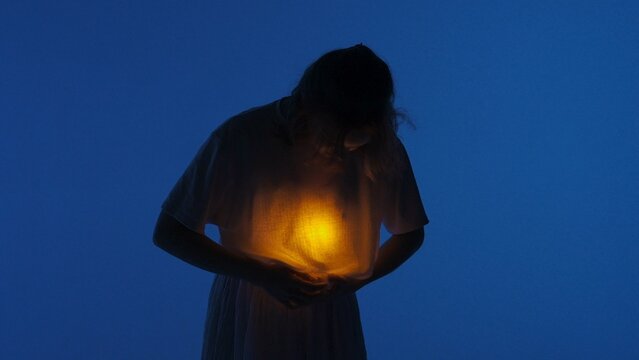 Female dancer performing in the studio. Young woman in dark studio touching warm light glowing inside the chest under her shirt.