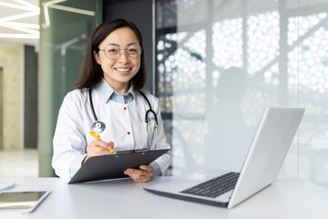 Portrait of a smiling Asian female doctor working in a hospital using a laptop, writing documents and case histories, consulting remotely and talking via video call.