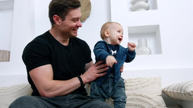 Father holds his lovely smiling baby boy standing beside on the sofa. Dad and son are at home.