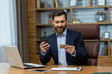 Young smiling and successful male businessman sitting in office at desk and using mobile phone and credit card.