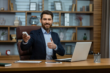 Portrait of a smiling and successful young businessman sitting at the office desk, turning on the...