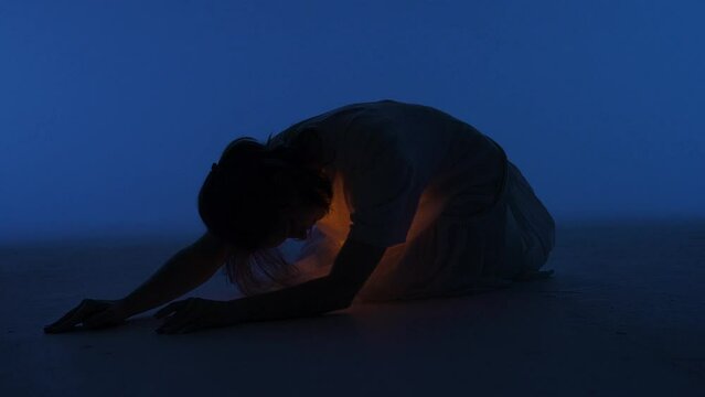 Female dancer performing in the studio. Young woman laying in dark studio with warm light glowing inside the chest under her shirt. Full shot 60 fps