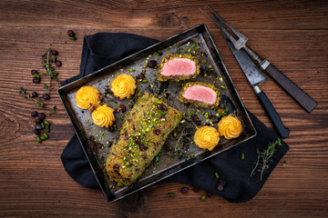 Gebratenes Mangalica Schweinefilet ummantelt mit zerkleinerten Pistazien, geriebenem Parmesan und gehackten Preiselbeeren serviert mit Prinzesskartoffeln als Draufsicht auf einem rustikalen schwarzen 