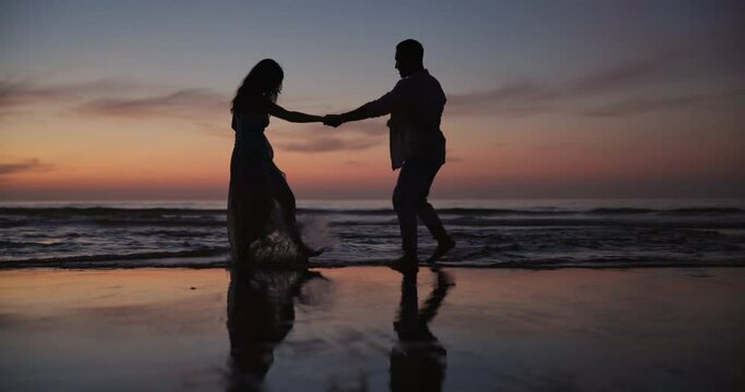 Couple, silhouette and dancing or holding hands on beach for bonding, water splash or fun outdoor evening together. Man and woman enjoying waves on ocean coast at night for holiday weekend in nature