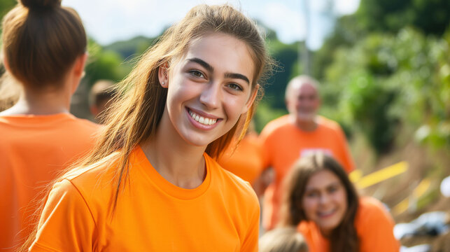 A cheerful young woman with a ponytail smiles brightly, wearing an orange volunteer t-shirt, with blurred group members in the background outdoors