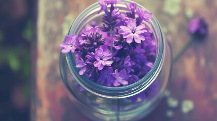  a jar filled with purple flowers sitting on top of a wooden table next to a green leafy plant on top of a wooden table.