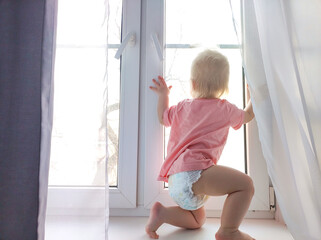 baby one year old stands on the windowsill, baby on the windowsill, trying to pull the window handle. Safety of children in the home © Виктория Чемаева